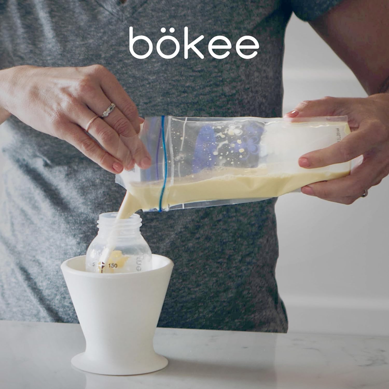 A person uses the bökee Baby Bottle Holder to effortlessly pour liquid from a zipper bag into a baby bottle with one hand, showcasing an accessible solution. The scene features the brand name "bökee" prominently above the person's hands.