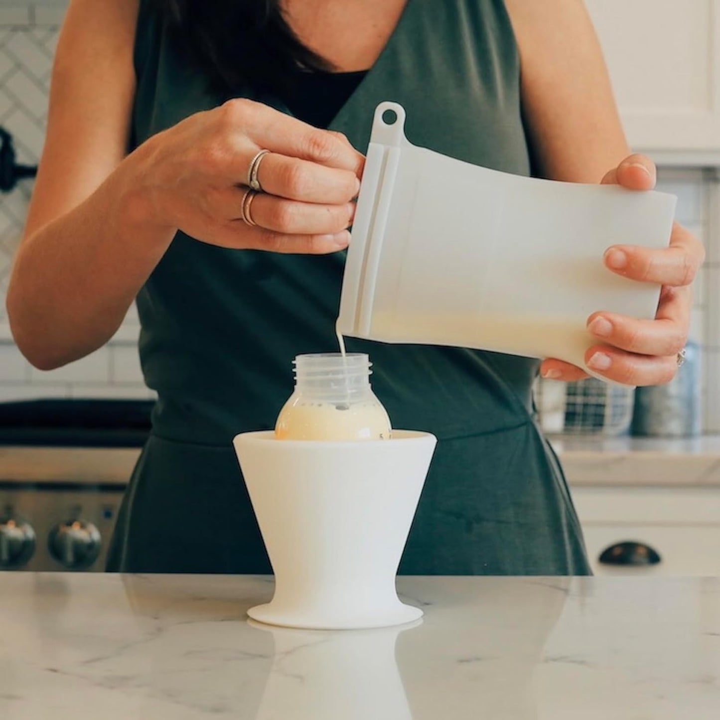 Using the bökee Baby Bottle Holder for one-handed bottle prep, a person pours milk from a BPA-free silicone bag into a baby's bottle with ease using a white funnel. Wearing a dark green sleeveless top, they are positioned by the stove against a backdrop of kitchen tiles.