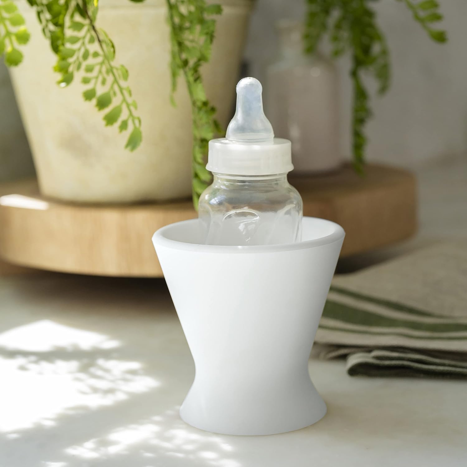 A bökee Baby Bottle Holder is positioned in a white cup on a sunlit surface. Nearby, green plants and a folded cloth accompany a light-colored wooden tray.