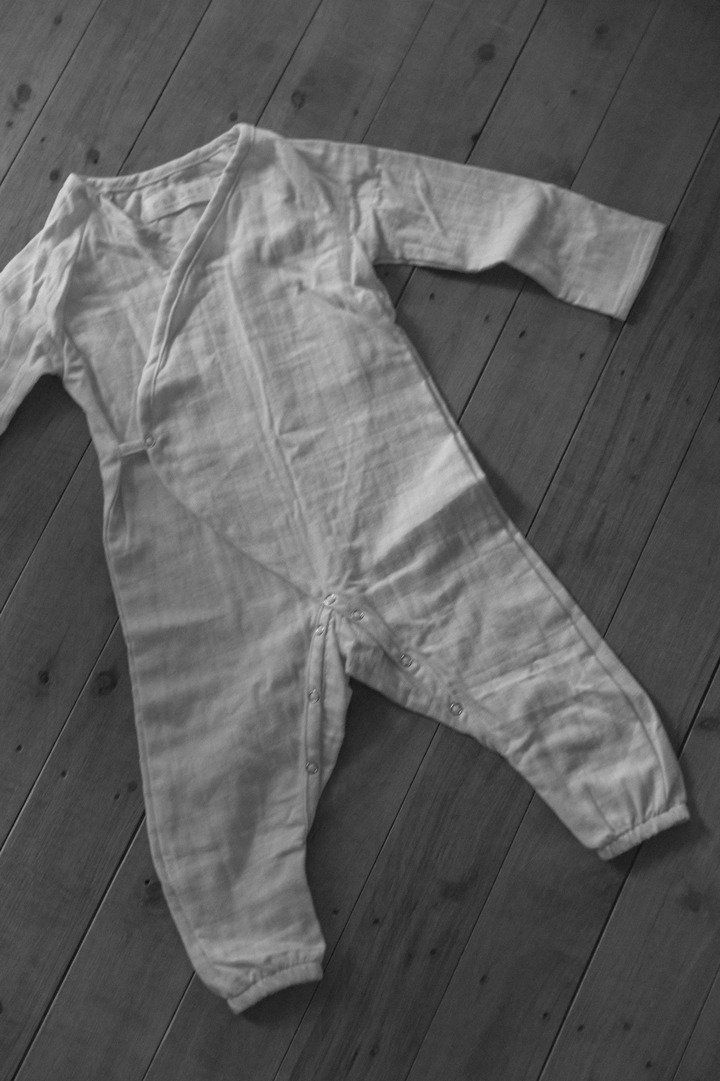 Image of the Après Organic Wrap Undyed baby onesie from cabane childrenswear, a classic piece from their babies' layette collection, laid flat on a wooden floor. Crafted from breathable fabric, this long-sleeved onesie features snap buttons along the legs and front for easy dressing.
