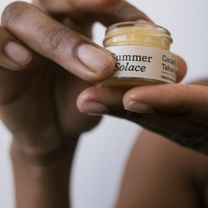 A person is holding a small jar of "Summer Solace Tallow" balm in their hands. The jar, labeled "Cacao Rose Cuticle, Lip, and Brow Balm - Regenerative Tallow™," is a handmade multipurpose moisturizer. The person's fingers are gently grasping the lid, preparing to open it.