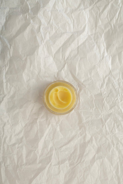 A small, round glass container filled with Summer Solace Tallow's Cacao Rose Cuticle, Lip, and Brow Balm - Regenerative Tallow™ sits atop a textured, crumpled white paper background, serving as a luxurious multipurpose moisturizer.