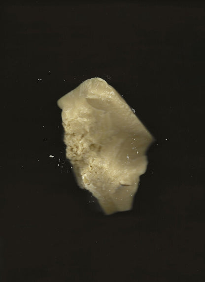 A partially bitten chunk of Cacao Rose Cuticle, Lip, and Brow Balm - Regenerative Tallow™ from Summer Solace Tallow, floating against a dark background. The off-white balm has a crumbly texture, with smaller pieces breaking off around it.