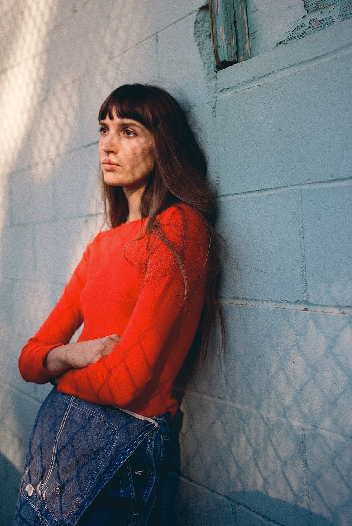Standing against a light blue brick wall with shadows from a chain-link fence, a person with long hair and a contemplative expression wears the Bebe Organic Long Sleeve by LA RELAXED and holds a denim jacket.