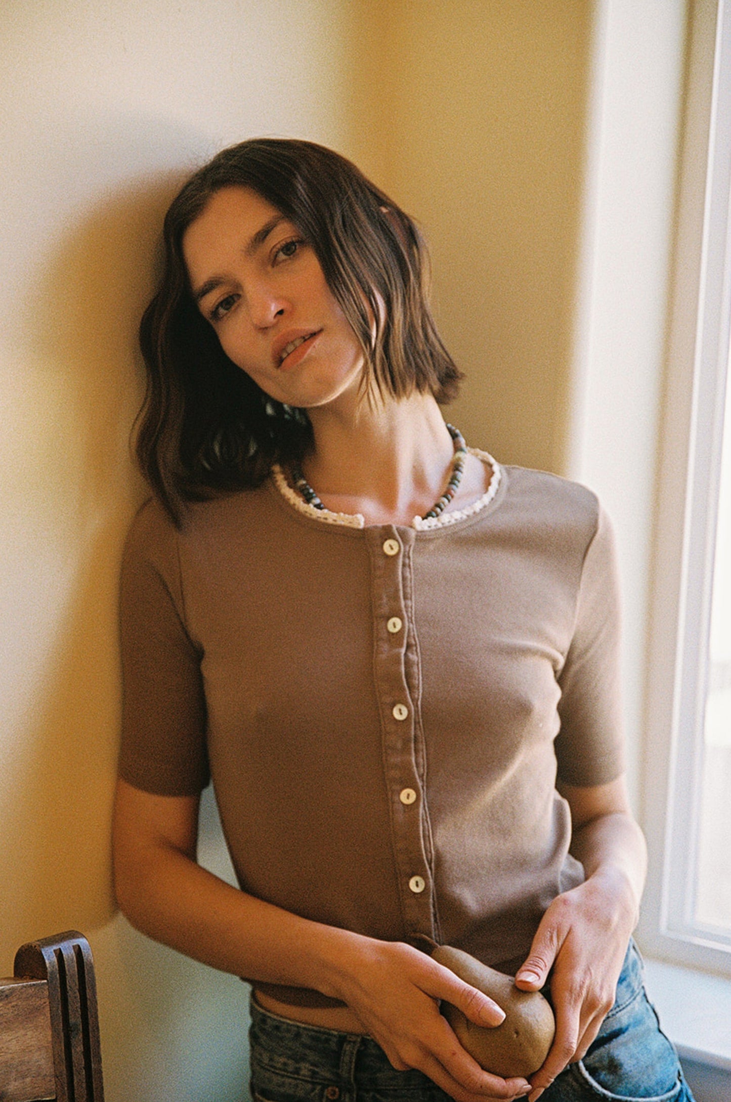 A person with shoulder-length dark hair leans against a wall in LA Relaxed's Short Sleeve Organic Cardi, complemented by a beaded necklace. They appear relaxed, holding an object while soft sunlight filters through a window beside them.