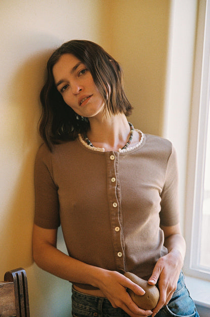 A person with shoulder-length dark hair leans against a wall in LA Relaxed's Short Sleeve Organic Cardi, complemented by a beaded necklace. They appear relaxed, holding an object while soft sunlight filters through a window beside them.