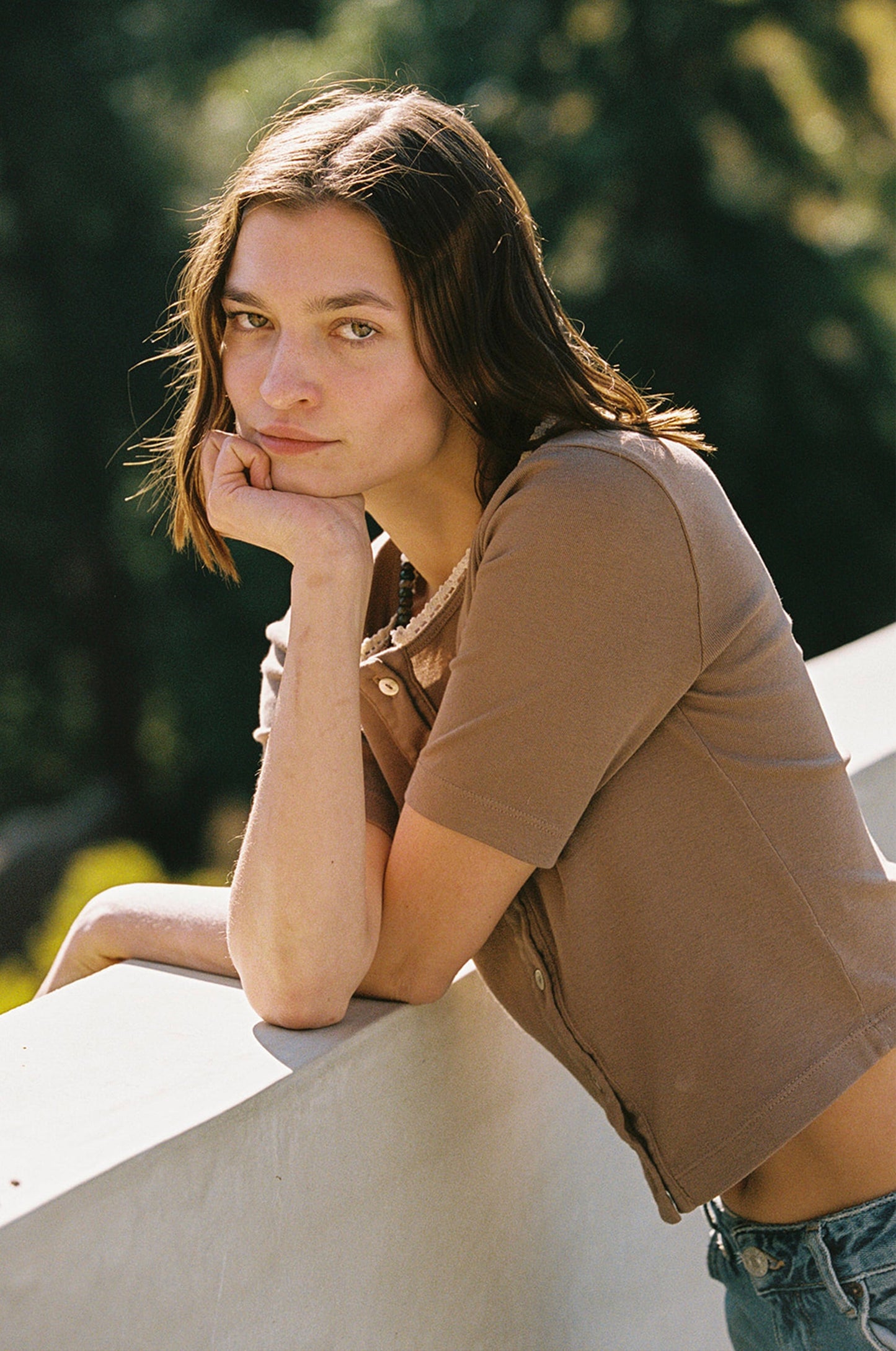 A young person with shoulder-length hair wears the LA Relaxed Short Sleeve Organic Cardi, which features delicate shell buttons. They lean thoughtfully on a white ledge outdoors, resting their chin on their hand against a softly blurred sunlit greenery backdrop.