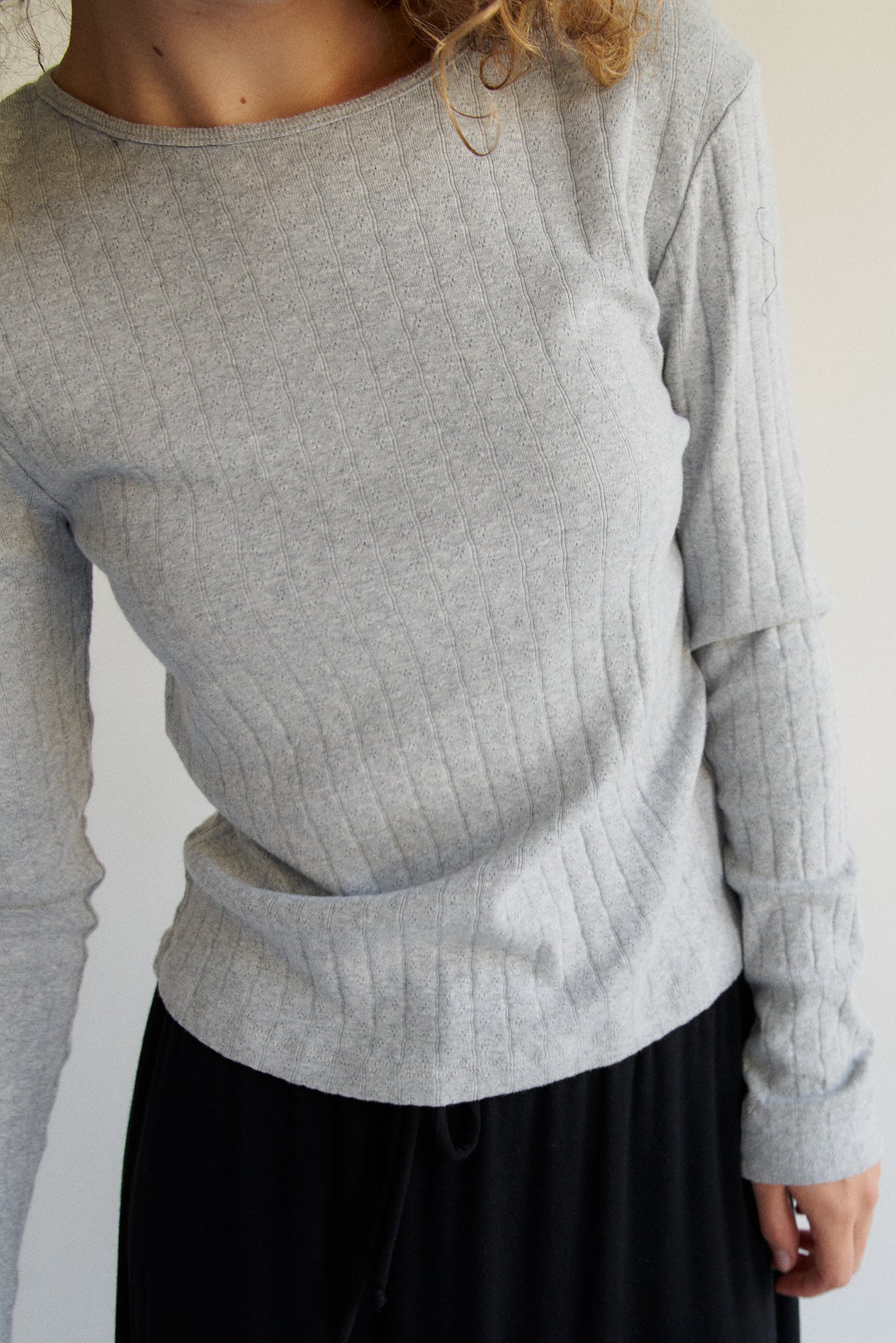An individual wears the LA RELAXED Organic Texture Long Sleeve sweater in light gray with a black skirt. Their face is hidden by curly hair against a plain, light-colored background.