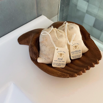 Two small drawstring bags labeled "rest" and "meditate" contain Honeybee Hippie's organic "meditate bath soak" salts, elegantly displayed in a wooden palm-shaped dish on a white surface. The bright setting, likely a bathroom, enhances the tranquil ambiance for meditation or foot soaks.