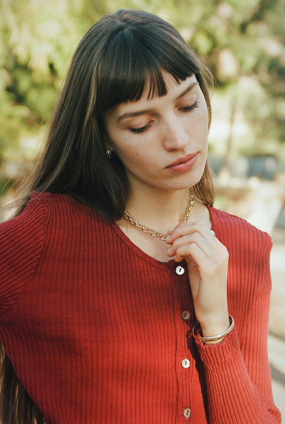 Cranberry-
 A young woman with long brown hair and bangs wears the LA RELAXED Pointelle Cardigan, featuring pearlescent buttons. She looks down thoughtfully, holding a gold necklace, against a softly blurred green outdoor background.