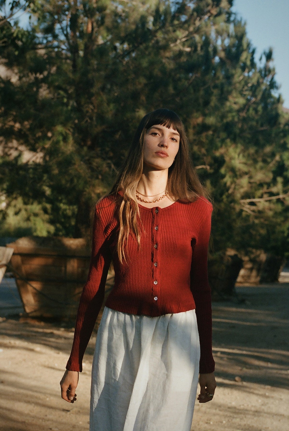 A woman with long brown hair stands outdoors in sunlight among trees, wearing the LA RELAXED Pointelle Cardigan with pearlescent buttons and a white skirt.