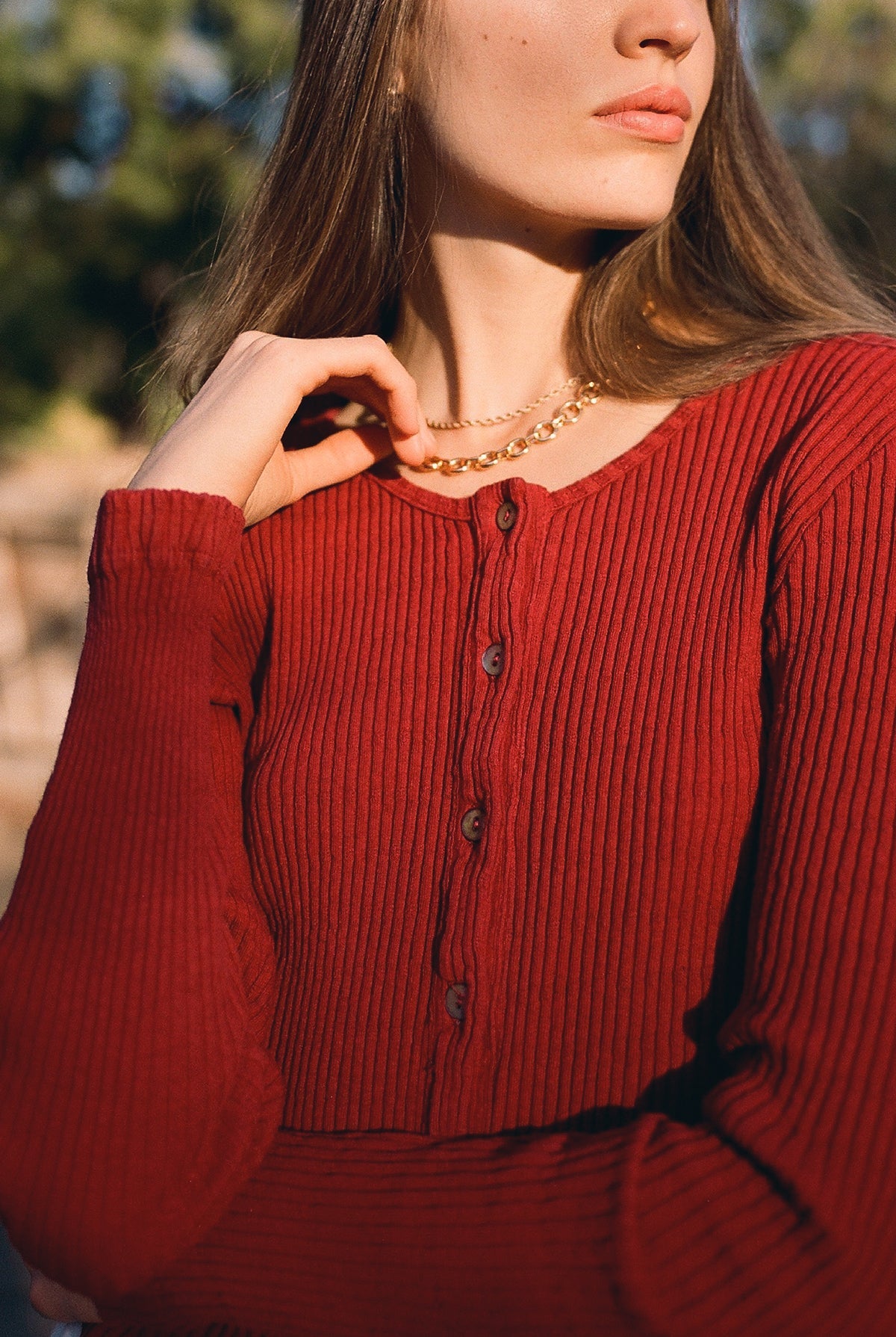 A woman stands outdoors wearing the LA RELAXED Pointelle Cardigan in red, featuring ribbed organic cotton and pearlescent buttons. She accessorizes with a gold chain necklace, her hand near her neck and her face partially visible as she looks to the side.