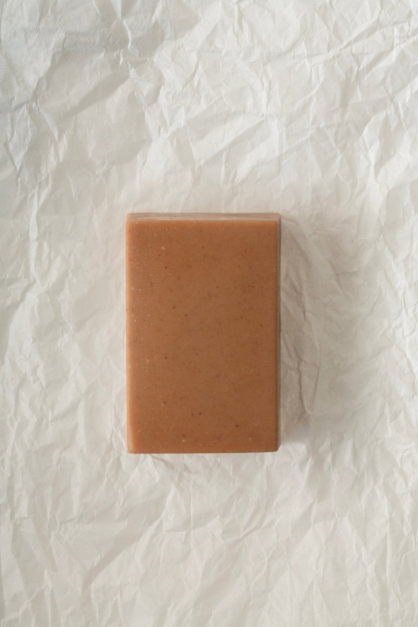 A rectangular bar of Desert Rose Red Clay Bar Soap - Regenerative Tallow™ by Summer Solace Tallow, with a light brown color, rests on crinkled white paper. Enriched with cacao butter, this hydrating soap has a smooth surface dotted with a few small speckles. The textured background contrasts beautifully with the clean lines of the soap bar.