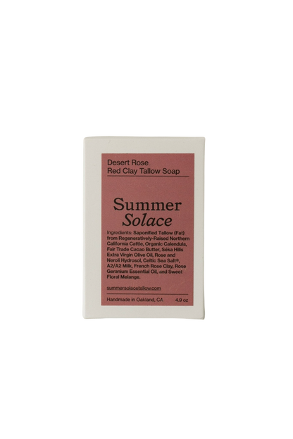 A rectangular bar of soap with a pink and white label reads "Desert Rose Red Clay Bar Soap - Regenerative Tallow™ by Summer Solace Tallow." The label lists ingredients such as hydrating tallow, cacao butter, various oils, botanicals, and rose essential oil. Handmade in Oakland, California, the soap weighs 4.9 oz.