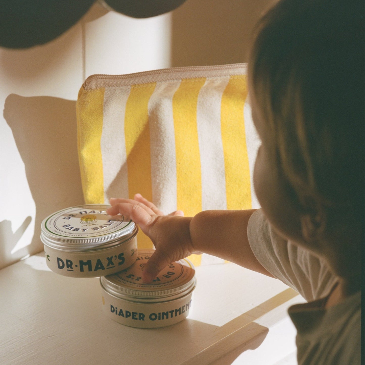 A child reaches for two tins from the Dr. Max’s Baby Essentials Bundle on a white surface, with a yellow and white striped pouch behind. Sunlight casts warm shadows, highlighting this gentle baby care moment.