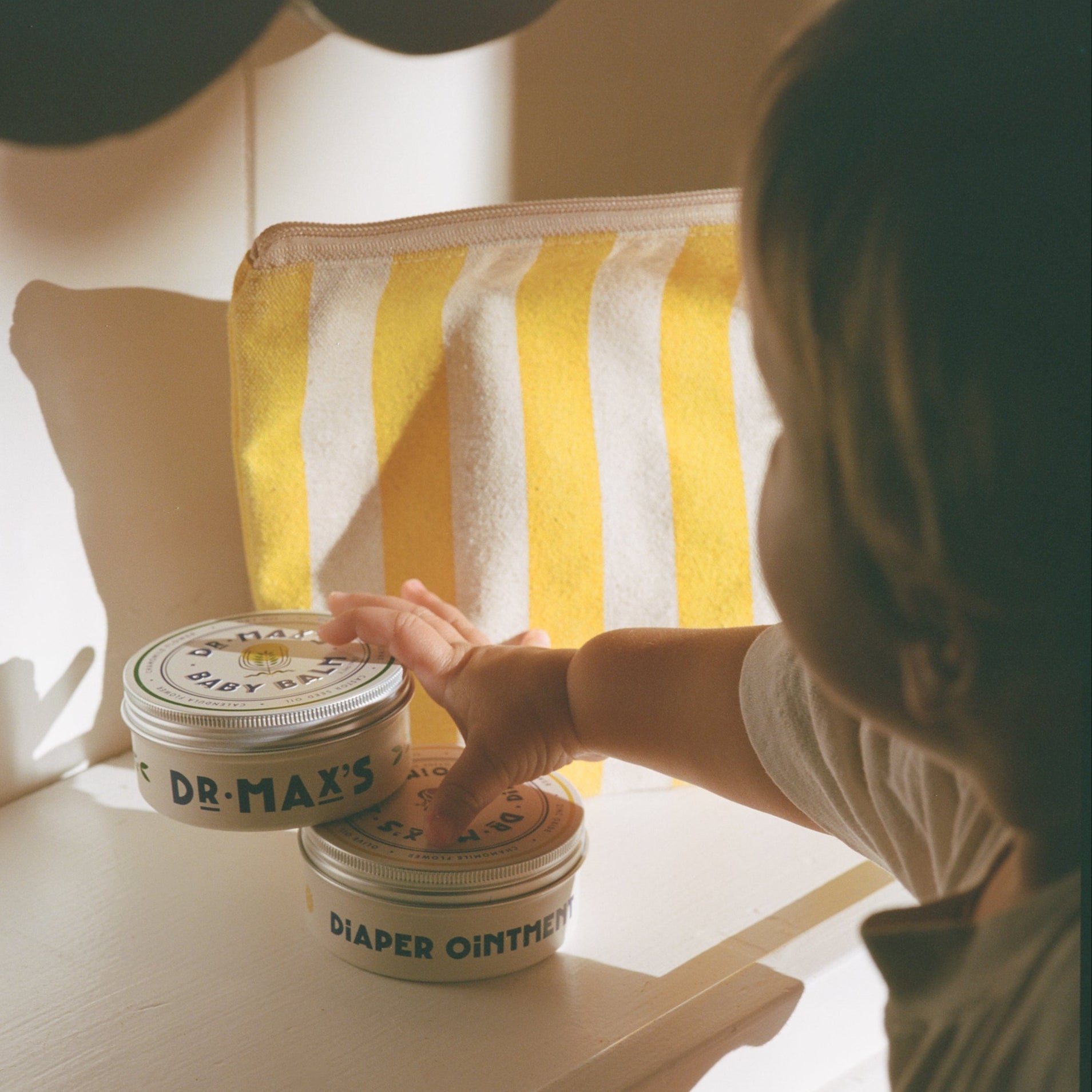 A child reaches for two tins from the Dr. Max’s Baby Essentials Bundle on a white surface, with a yellow and white striped pouch behind. Sunlight casts warm shadows, highlighting this gentle baby care moment.