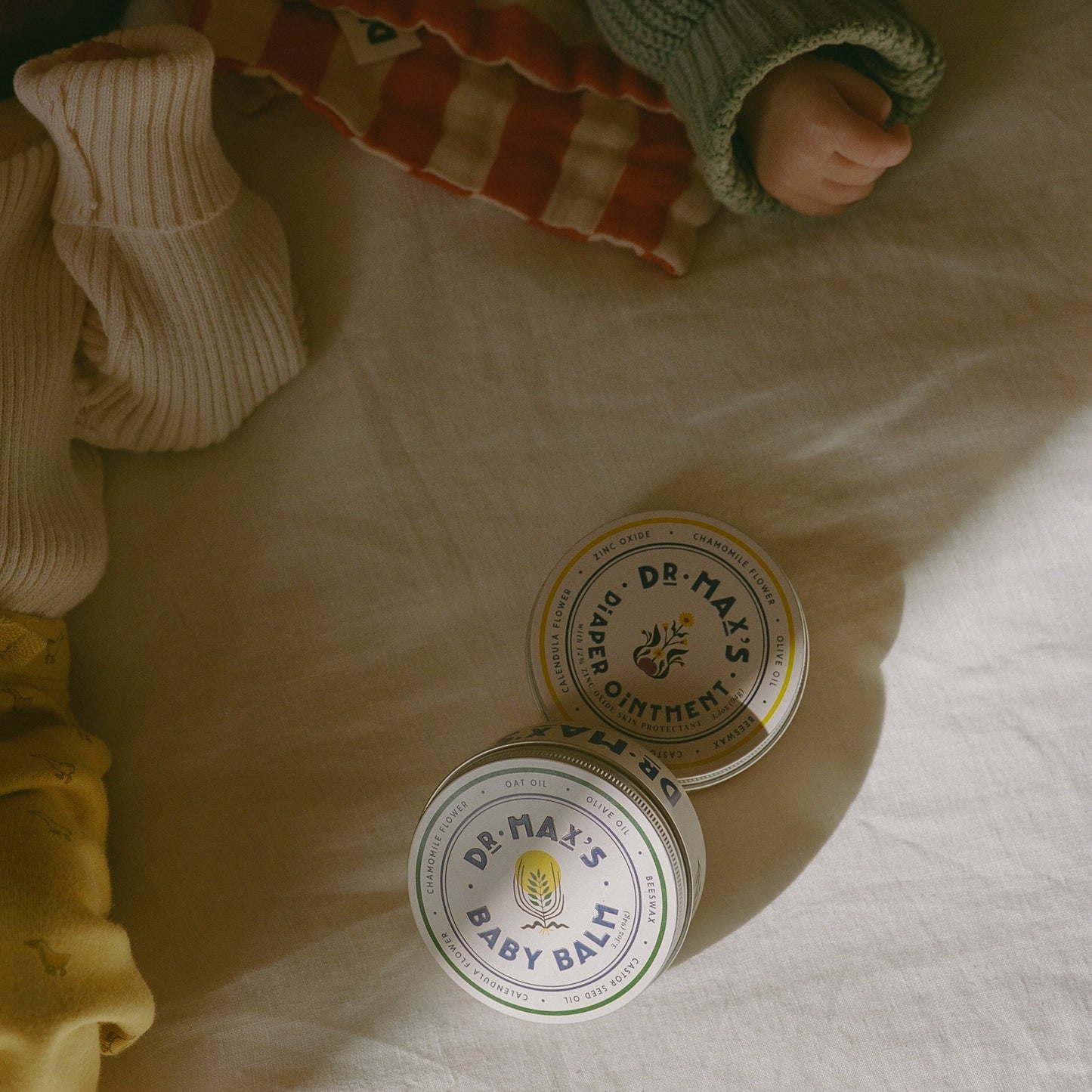 A Newborn Gift Box from Dr Maxs rests on cream fabric next to a baby in yellow pants and a green sweater, showcasing gentle care products for delicate skin.