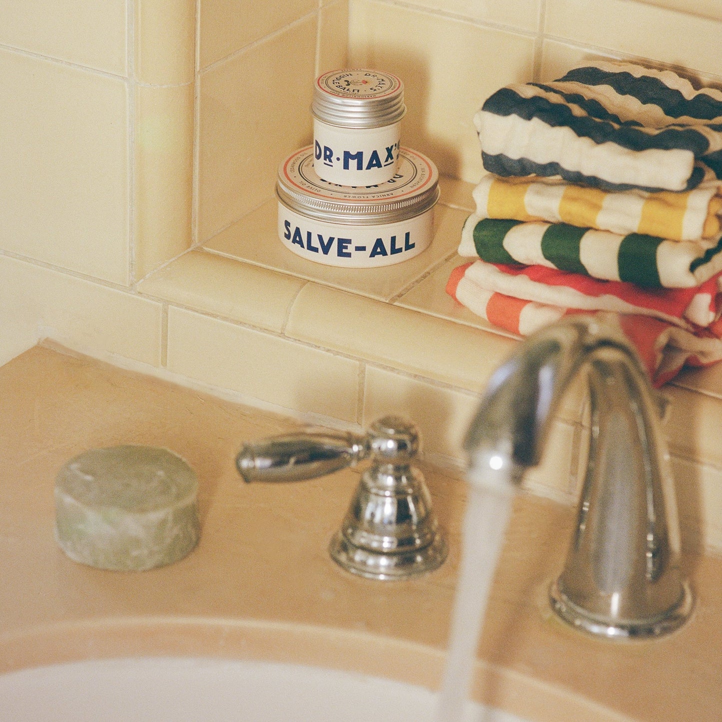 A bathroom countertop displays Dr. Max's Adult Essentials Bundle: a round green Olive Oil Soap bar, stacked striped towels, and two tins labeled "Dr. Max's" and "SALVE-ALL," beside a silver faucet running water on a tiled shelf.