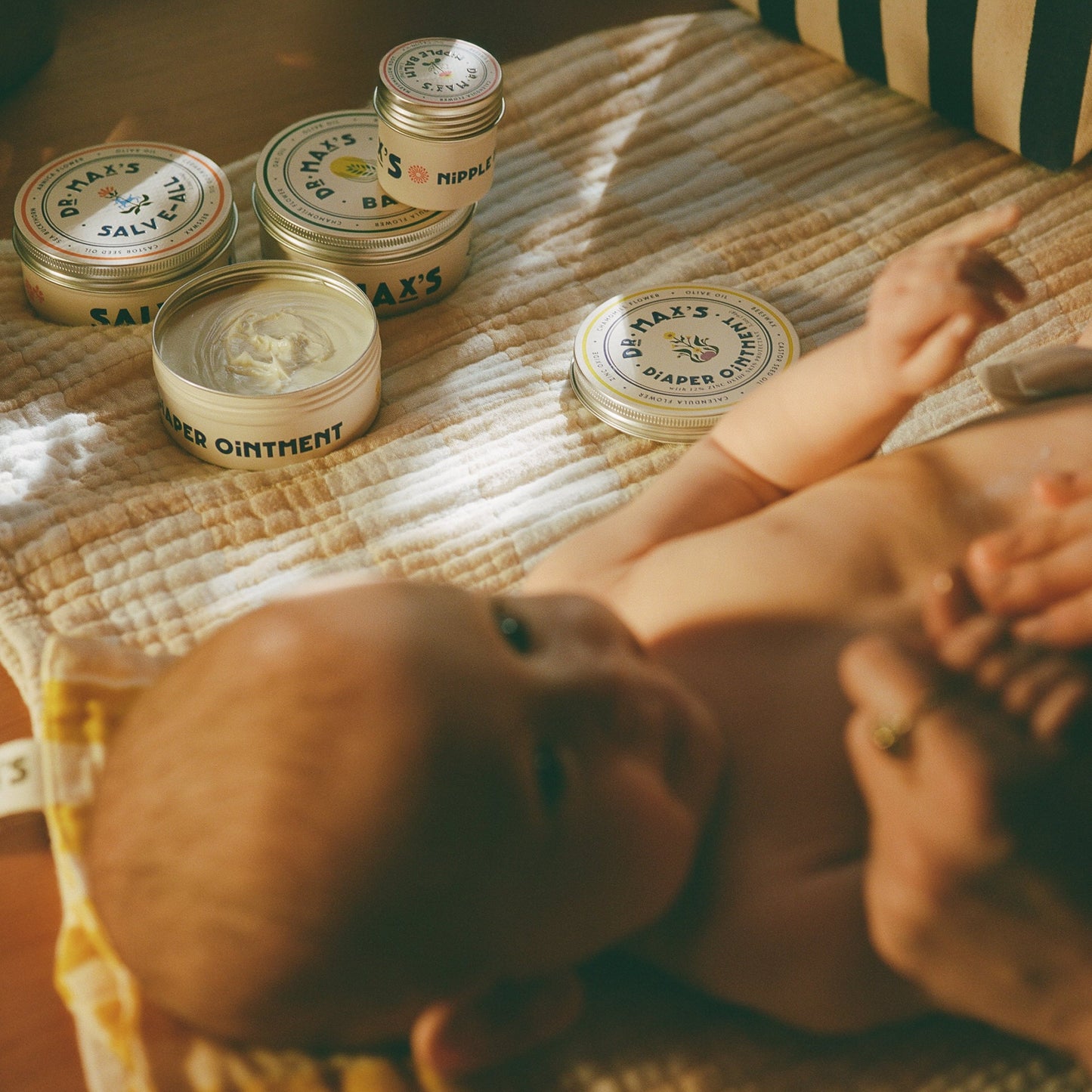 A baby rests on a mat, surrounded by Dr Maxs New Parent Gift Box essentials—Baby Balm Diaper Ointment, salves—and the cozy Dr. Animals children’s book, all in soft, warm lighting for gentle comfort and storytime.