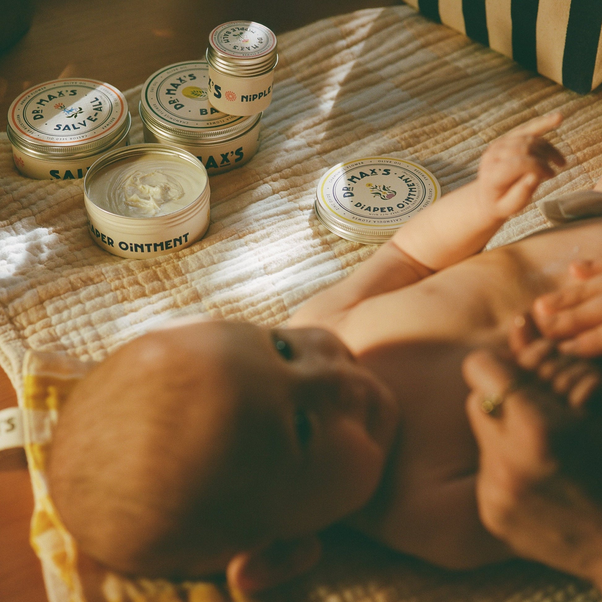 A baby rests on a mat, surrounded by Dr Maxs New Parent Gift Box essentials—Baby Balm Diaper Ointment, salves—and the cozy Dr. Animals children’s book, all in soft, warm lighting for gentle comfort and storytime.
