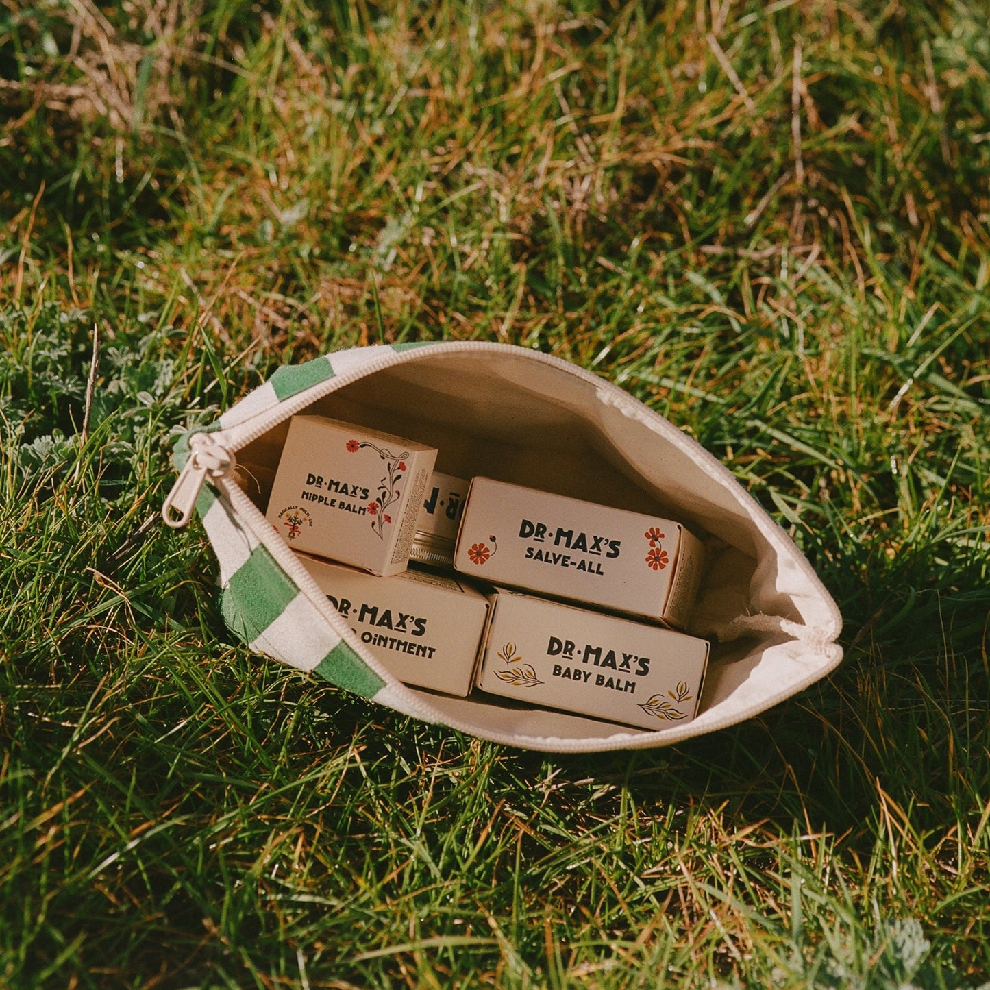 A small green-and-white striped zippered pouch sits open on grass, displaying Dr Maxs’ New Parent Gift Box with Baby Balm Diaper Ointment and other soothing ointments and balms inside.