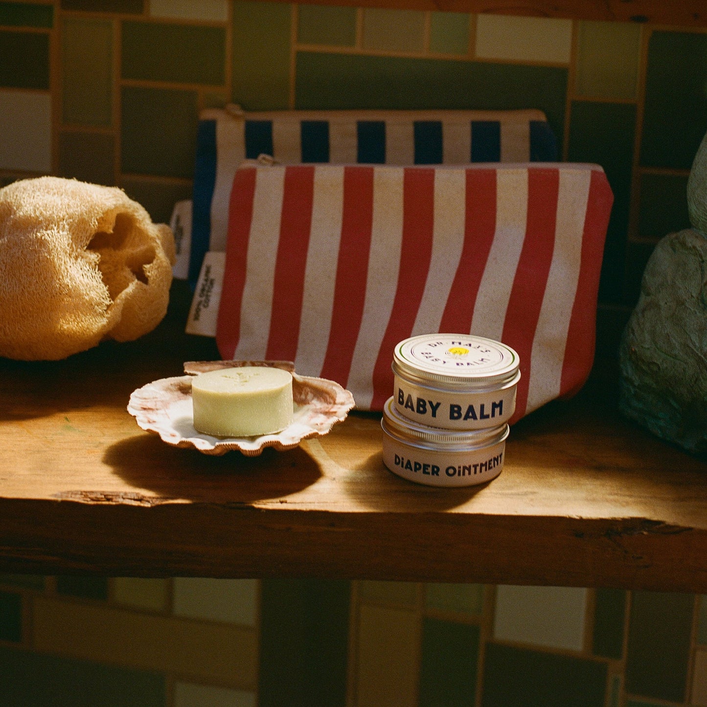 A wooden shelf holds items from the Dr Maxs Newborn Gift Box—including a striped pouch, yellow sponge, Olive Oil Soap on a dish, and Baby Balm Diaper Ointment tin—displayed against a tiled wall.