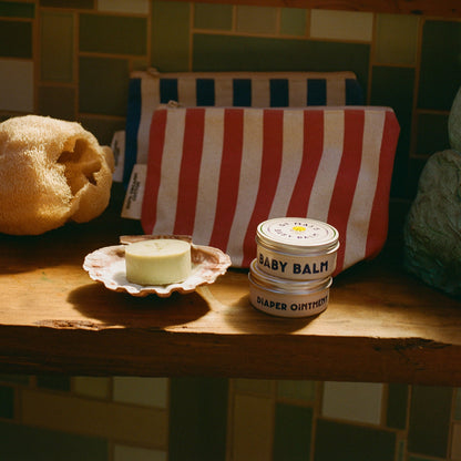A wooden shelf holds items from the Dr Maxs Newborn Gift Box—including a striped pouch, yellow sponge, Olive Oil Soap on a dish, and Baby Balm Diaper Ointment tin—displayed against a tiled wall.