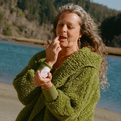 An older woman with long, wavy gray hair stands by a river, using her finger to apply Dr. Max's Adult Essentials Bundle to her lips. Trees and water can be seen in the background.
