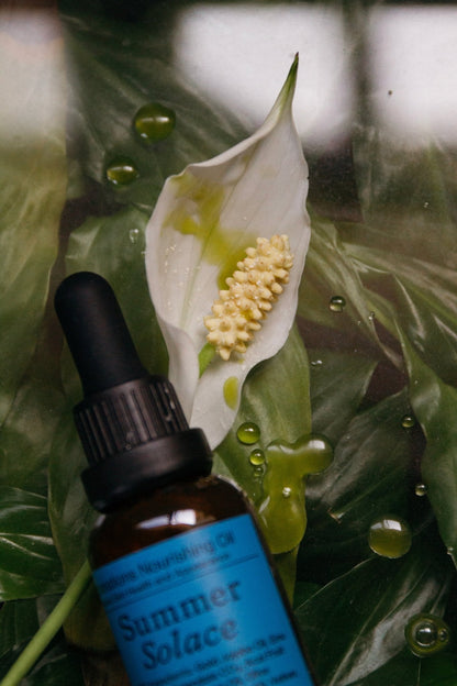A brown glass dropper bottle labeled "Summer Solace Tallow" sits on lush green leaves by a white peace lily, with drops of Emanations Profound Face Oil—Deeply Nourishing, featuring champa, tuberose, white ginger lily, and rich tallow—scattered nearby.