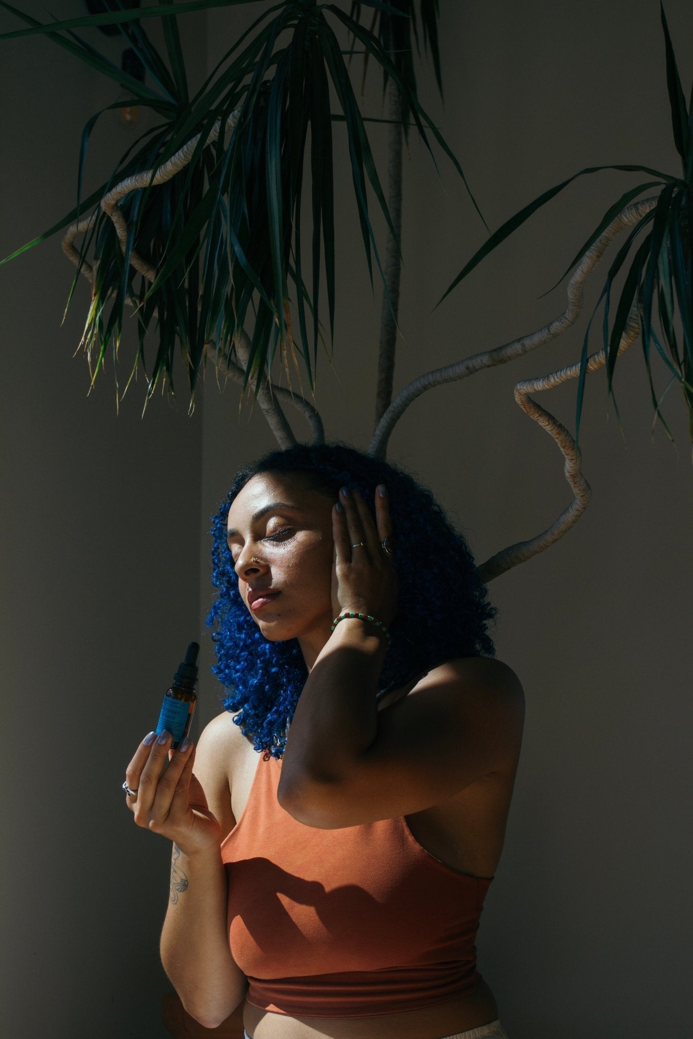 A woman with blue curly hair in a rust tank top stands in sunlight, applying Summer Solace Tallow’s Emanations Profound Face Oil – Deeply Nourishing (Champa, Tuberose & White Ginger Lily) with plant branches arching above her.