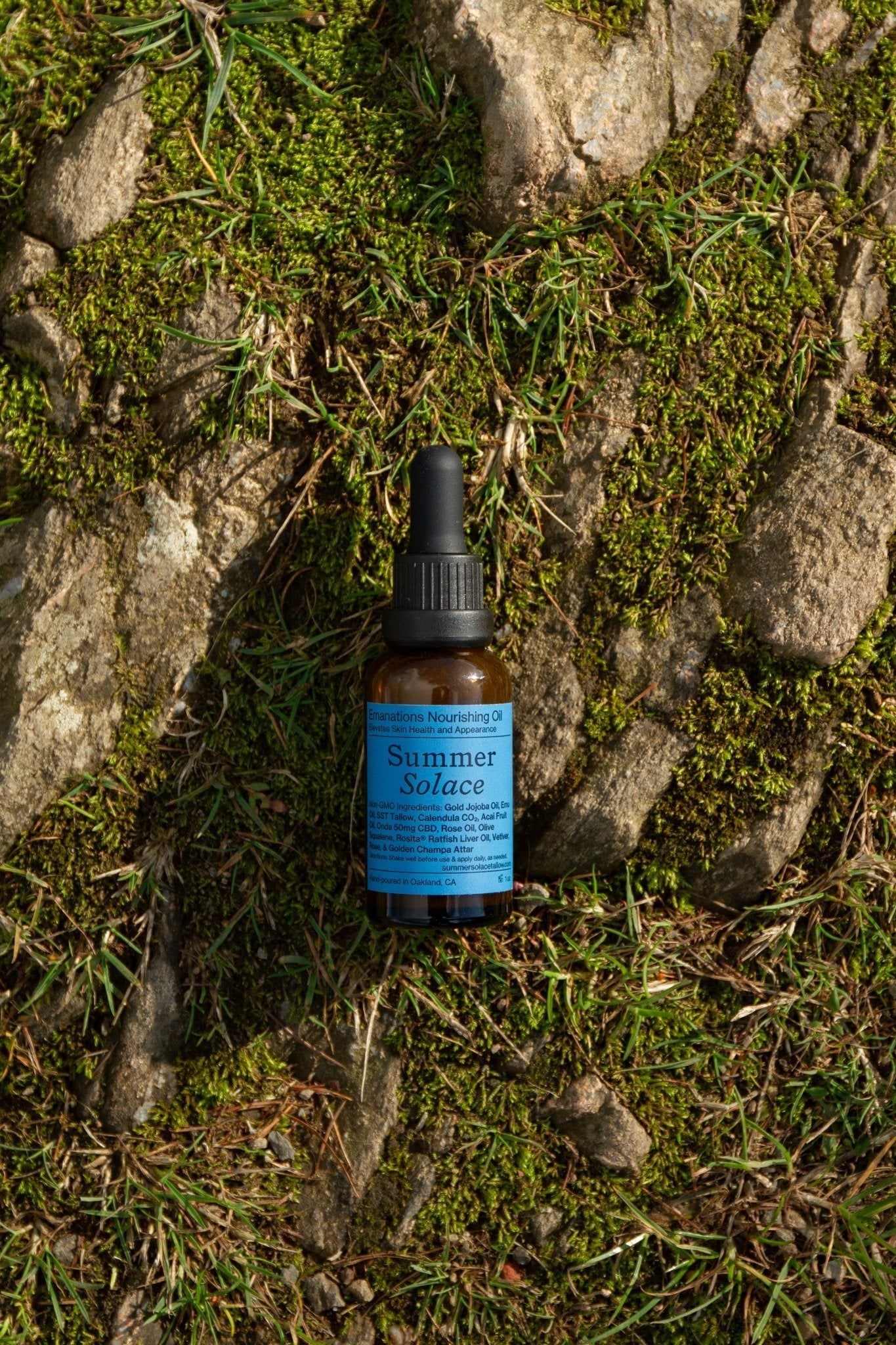 A small brown dropper bottle labeled "Summer Solace Tallow"—Emanations Profound Face Oil with Champa, Tuberose & White Ginger Lily—rests on mossy, sunlit ground among patches of grass and exposed stone.