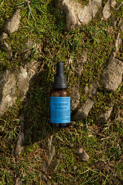 A small brown dropper bottle labeled "Summer Solace Tallow"—Emanations Profound Face Oil with Champa, Tuberose & White Ginger Lily—rests on mossy, sunlit ground among patches of grass and exposed stone.