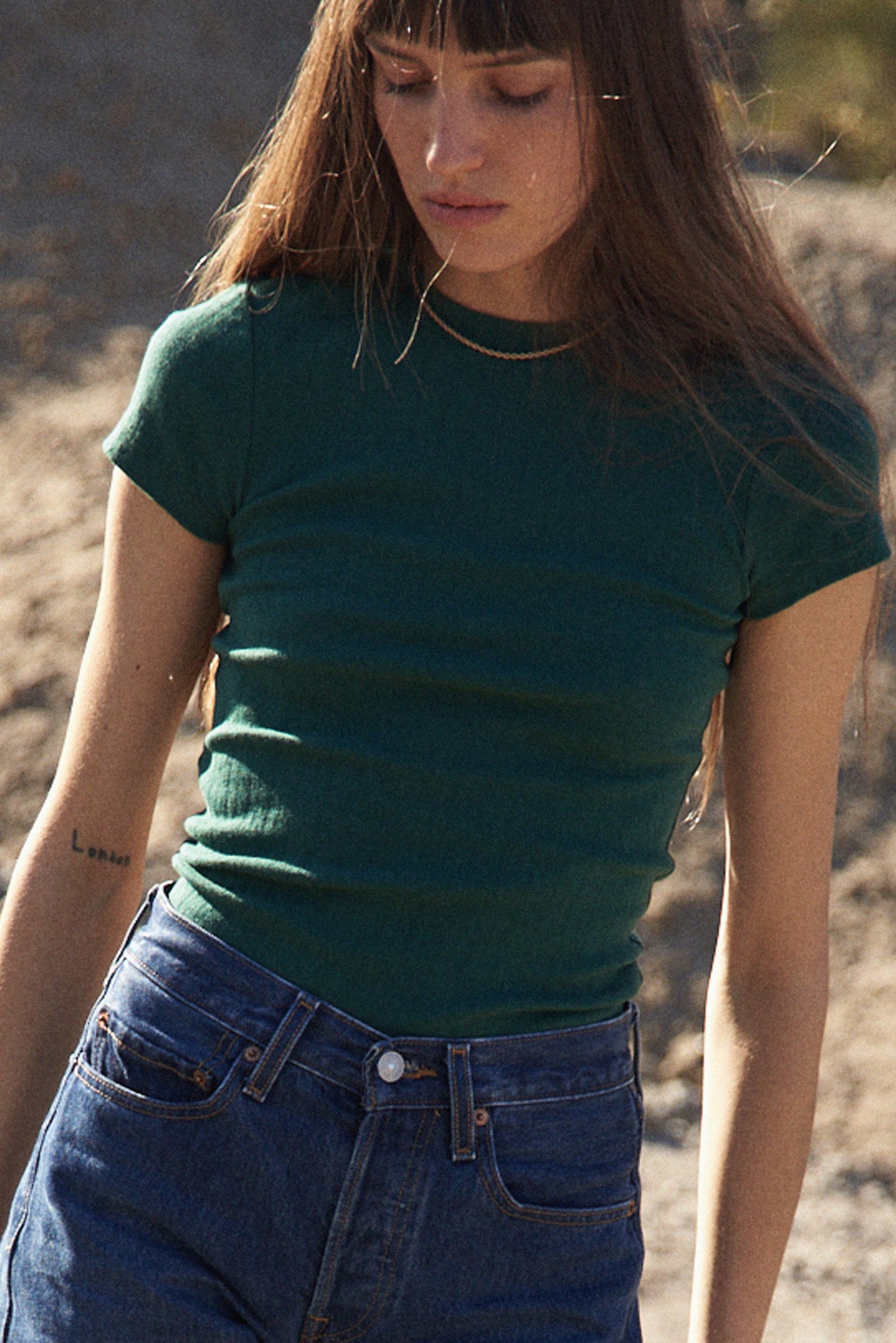 A person with long brown hair and bangs, clad in a green Organic Texture Tee by LA RELAXED and blue jeans, stands outdoors gazing downward. A tattoo adorns their left arm against a sunlit sandy backdrop.