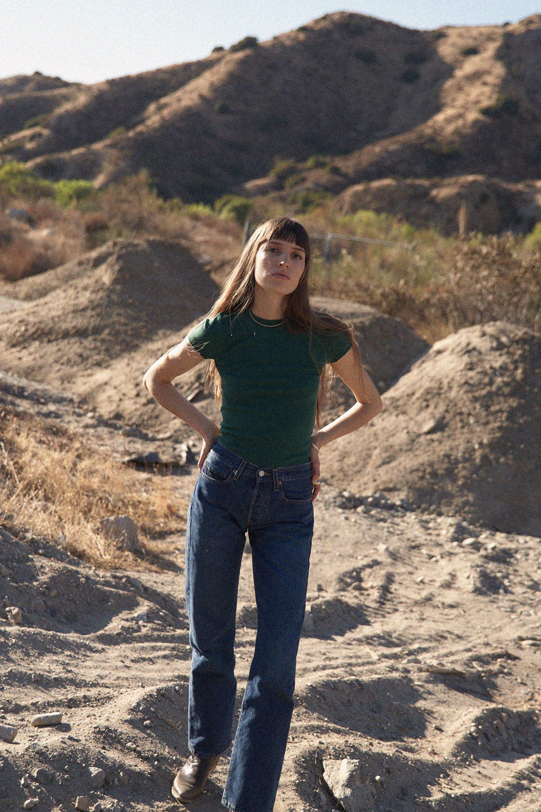 A woman wearing the LA RELAXED Organic Texture Tee and jeans stands on a rocky, barren landscape with hills in the background under a clear blue sky.