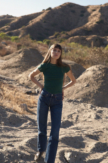 A woman wearing the LA RELAXED Organic Texture Tee and jeans stands on a rocky, barren landscape with hills in the background under a clear blue sky.