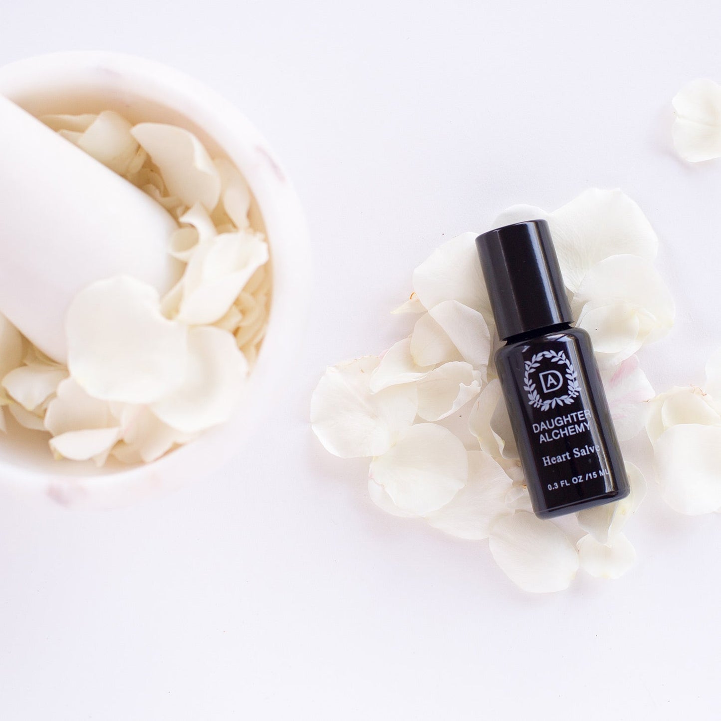 A Heart Salve® Botanical Nurturing Oil roller bottle from Daughter Alchemy is placed on a white surface surrounded by white flower petals. To the left, a mortar filled with similar white petals is partially visible, highlighting the organic ingredients used in this nurturing oil.