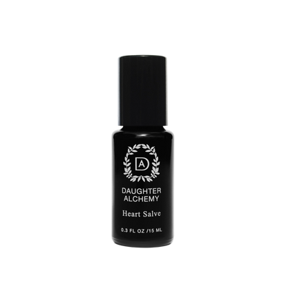 A small black bottle of Heart Salve® Botanical Nurturing Oil by Daughter Alchemy, crafted with organic ingredients, is displayed against a black background. The label features a white logo with a wreath design around the initials "DA" above the product name and indicates the bottle contains 0.3 fl oz (15 ml) of nurturing oil salve.