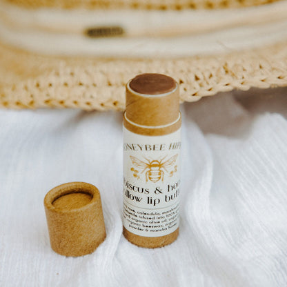 A natural lip butter infused with organic herbs, the Honeybee Hippie hibiscus & honey tallow comes in a cardboard tube on white cloth. The cap is beside the open tube, and a straw hat is blurred in the background.