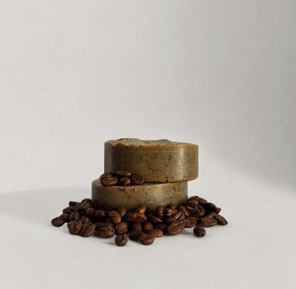 Two round bars of Wild Mamas' Wild Bison Tallow Deep Exfoliating Soap Scrub—Shilajit + Decaffeinated Coffee Bean—are stacked with coffee beans scattered around, highlighting the regenerative ingredients, against a plain white background.