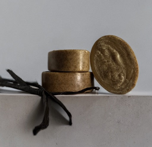 Three round, brown Wild Mamas Wild Bison Tallow Gentle Exfoliating Soap Scrub — Madagascar Vanilla Bean + Manuka Honey bars are stacked on a white surface with two dried vanilla pods beside them, set against a light, plain background.