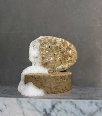 Two oval bars of Wild Mamas’ Wild Bison Tallow Deep Exfoliating Soap Scrub—Shilajit + Decaffeinated Coffee Bean—are stacked on marble, with the top bar foamy and white against a dark, smooth wall background.
