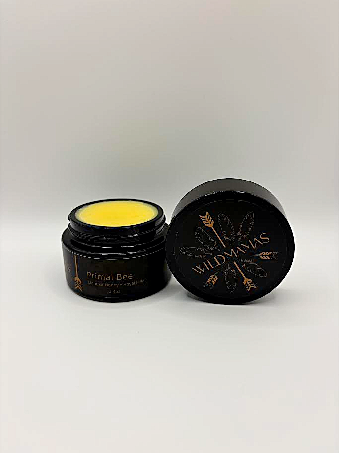 An open jar of Wild Mamas Wildly Nourishing Bison Tallow Balm - Primal Bee (Manuka & Royal Jelly) reveals yellow balm inside. The round lid, set beside the jar, features the Wild Mamas logo with feather and gold accents.