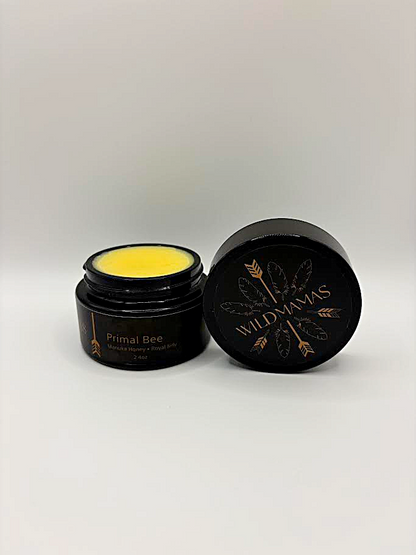 An open jar of Wild Mamas Wildly Nourishing Bison Tallow Balm - Primal Bee (Manuka & Royal Jelly) reveals yellow balm inside. The round lid, set beside the jar, features the Wild Mamas logo with feather and gold accents.