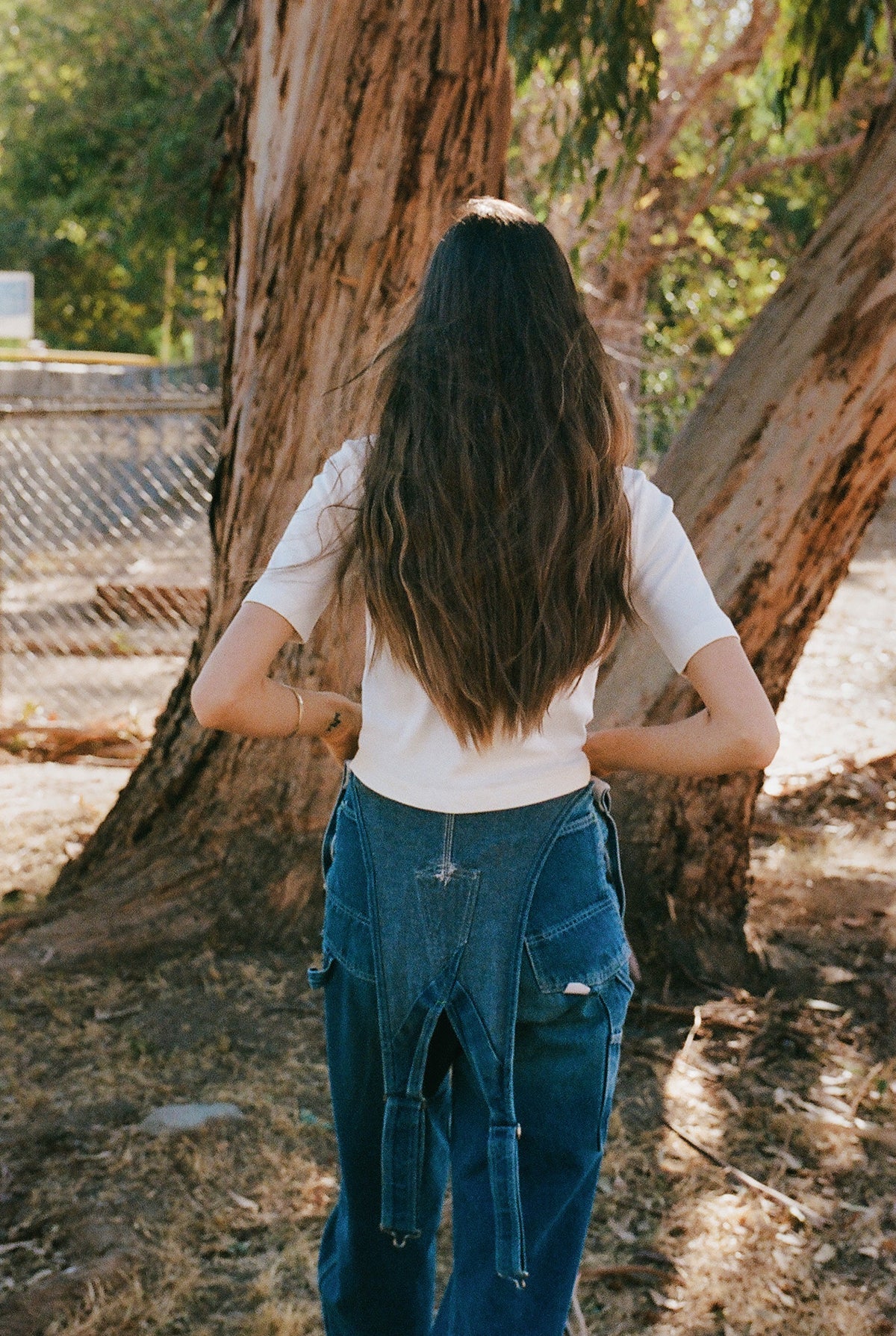 A person with long brown hair stands outdoors, back to the camera, wearing an LA Relaxed Short Sleeve Organic Cardi. They are near large tree trunks and a chain-link fence, with sunlight gently filtering through the trees.