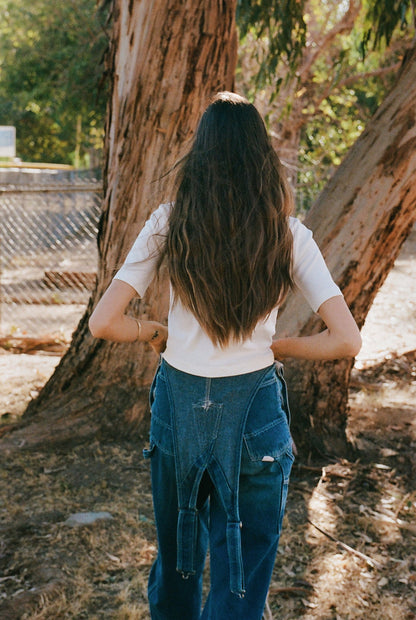 A person with long brown hair stands outdoors, back to the camera, wearing an LA Relaxed Short Sleeve Organic Cardi. They are near large tree trunks and a chain-link fence, with sunlight gently filtering through the trees.