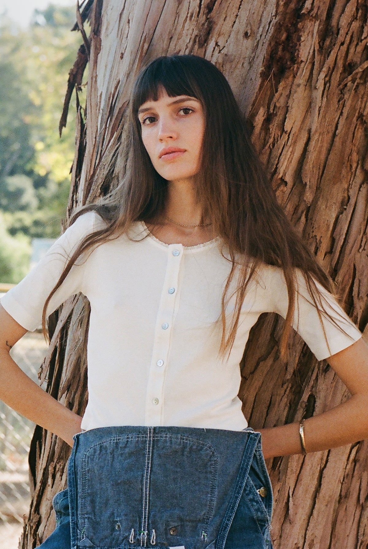 A person with long brown hair in a LA Relaxed Short Sleeve Organic Cardi and blue overalls stands against a tree. The background is blurred with greenery. They have a calm expression, and one hand is tucked into a pocket.