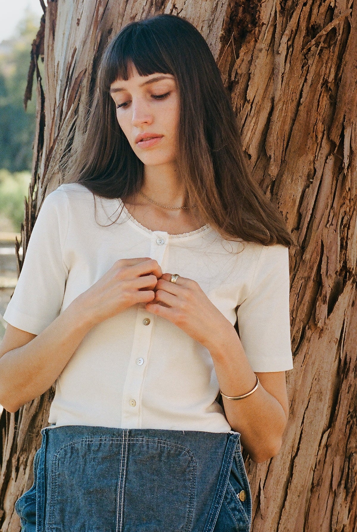 A woman with long brown hair stands against a tree, wearing a white LA Relaxed Short Sleeve Organic Cardi and denim pants. She looks down calmly, gently holding the collar of her shirt.
