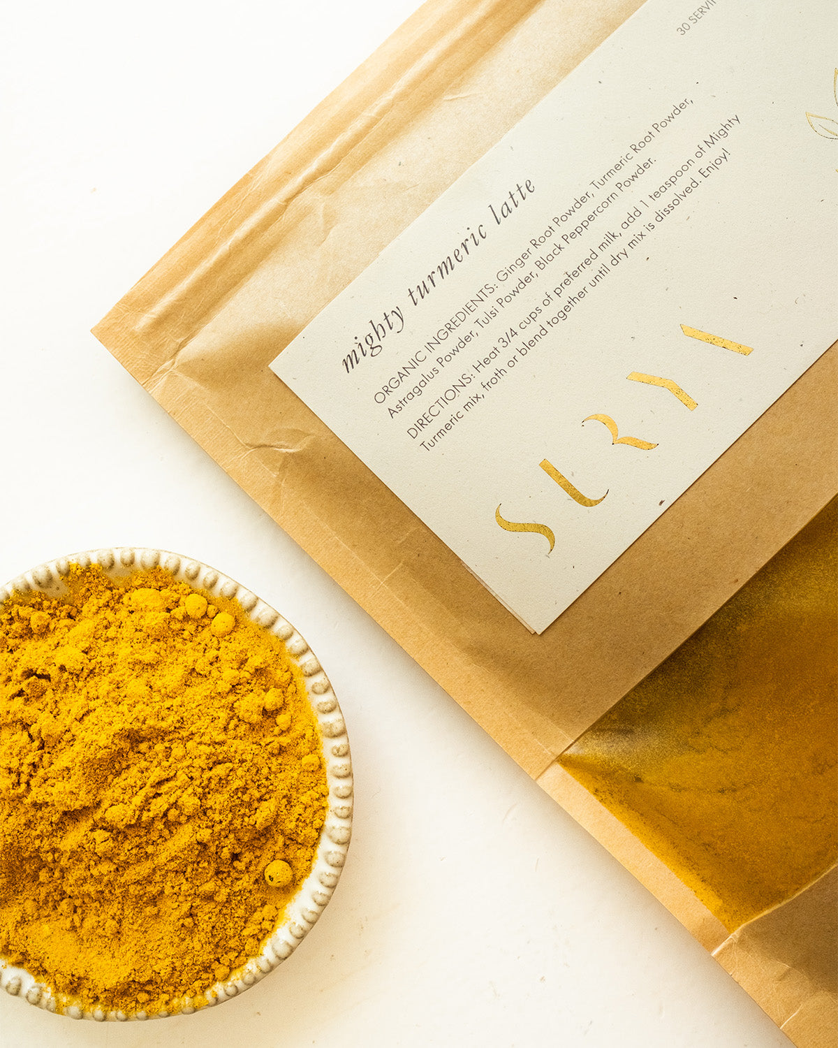 A brown paper packet labeled "Mighty Turmeric Latte," prominently featuring the brand name "Surya" in gold letters, is positioned beside a small white bowl brimming with vibrant yellow turmeric powder. The ingredients and instructions on the packet are partially visible, making it ideal for incorporating Ayurvedic latte mixes into your wellness routine.