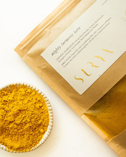 A brown paper packet labeled "Mighty Turmeric Latte," prominently featuring the brand name "Surya" in gold letters, is positioned beside a small white bowl brimming with vibrant yellow turmeric powder. The ingredients and instructions on the packet are partially visible, making it ideal for incorporating Ayurvedic latte mixes into your wellness routine.