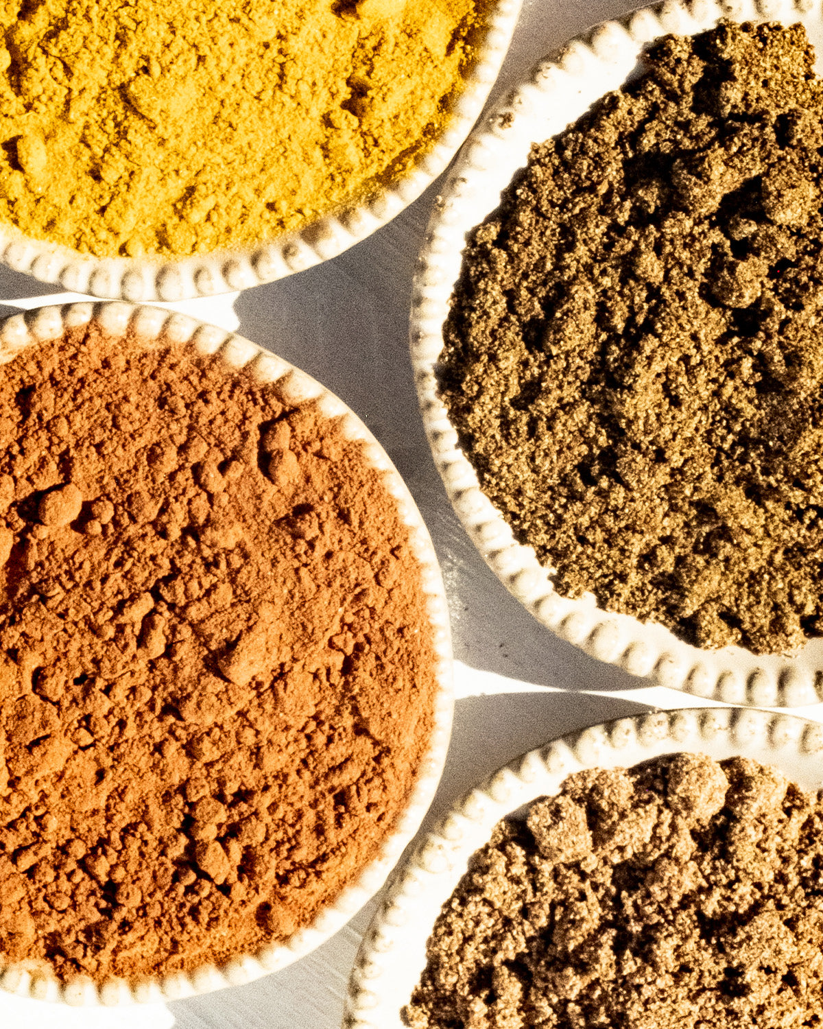 An overhead view of four small, round, white ceramic bowls, each filled with different colored powdered spices. The top-left bowl contains a bright yellow spice often used in Surya's Mighty Turmeric Latte mixes; the top-right a greenish-brown spice; the bottom-left a reddish-brown spice known for its anti-inflammatory properties; and the bottom-right a light brown spice.